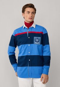 Kleurrijk overhemd met knopen en horizontale strepen in lichtblauw, marineblauw en rood. Bevat een borstzak met borduursel en een witte kraag.