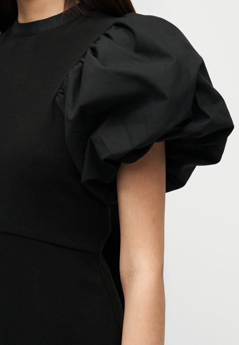 Abito nero con grandi maniche a sbuffo arricciate. Il corpo è aderente, realizzato in tessuto elasticizzato, con scollo tondo e texture liscia.