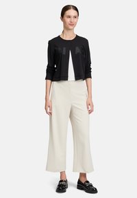 Kobieta w czarnym, krótkim kardiganie z napisem, białych, szerokich spodniaczkach za kostkę i czarnych, masywnych loafersach, stojąca na białym tle.