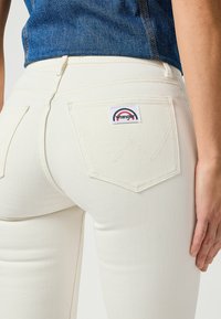 Valkoiset denim-farkut, joissa on tiukka malli, takataskut ja Wrangler-logo-patch. Sileä pinta näkyvillä ompeluyksityiskohdilla.