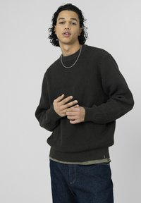 Pull en maille texturée gris foncé avec poignets et ourlet côtelés, porté sur une chemise vert clair, associé à un jean denim foncé. Collier chaîne argentée visible.