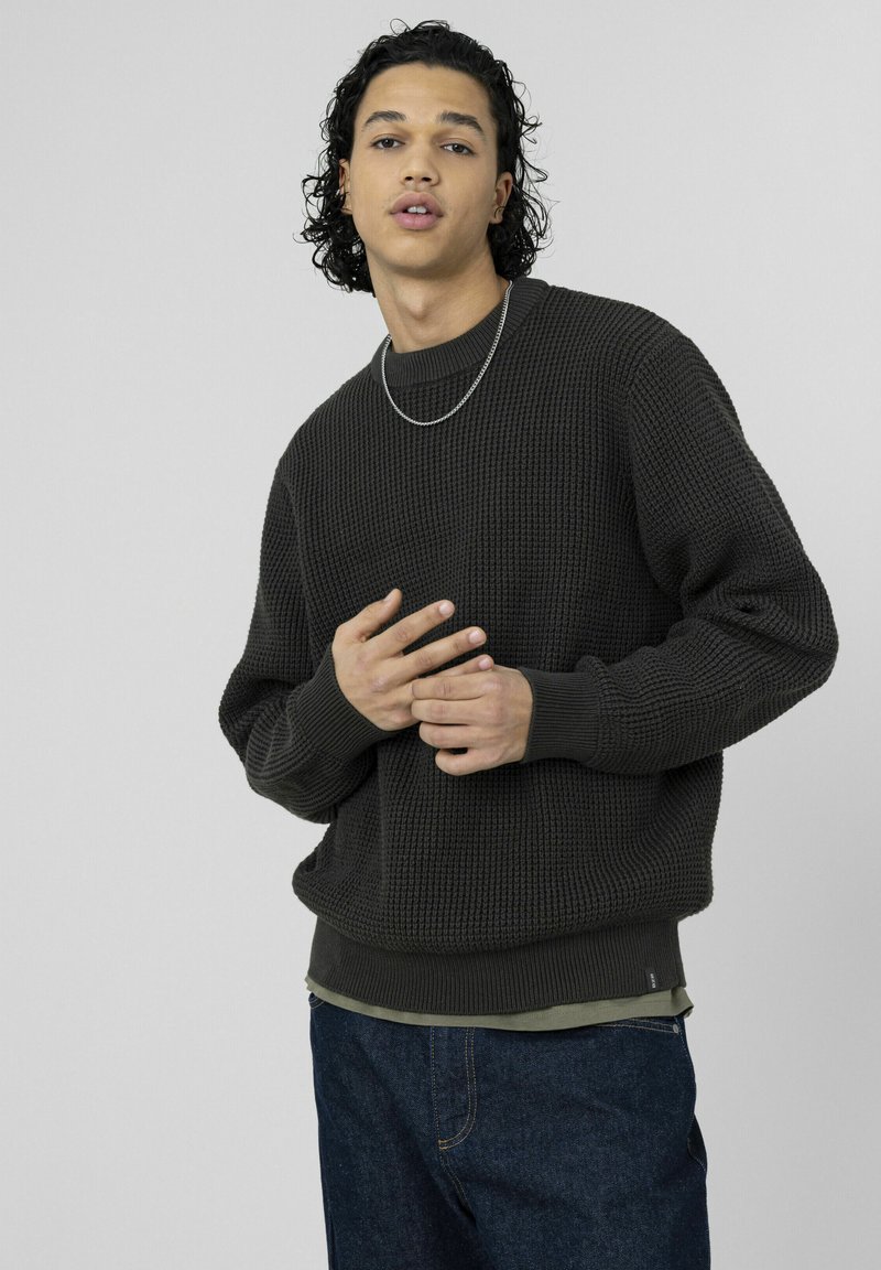 Pull en maille texturée gris foncé avec poignets et ourlet côtelés, porté sur une chemise vert clair, associé à un jean denim foncé. Collier chaîne argentée visible.