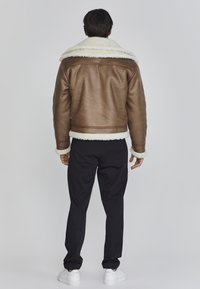 Chaqueta de cuero marrón con un cuello de borrego blanco, puños acanalados y dobladillo. LLevada con pantalones negros y zapatillas blancas.