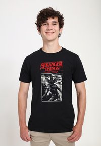 Camiseta de algodón negra con un audaz texto rojo "STRANGER THINGS" encima de un gráfico vintage en blanco y negro de una criatura.
