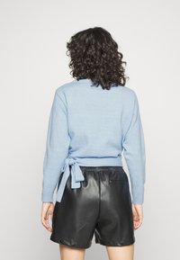Suéter azul claro de manga larga con un cierre atado en la parte posterior, combinado con shorts de cuero negros que tienen detalles de botones a presión y una cinturilla elástica.