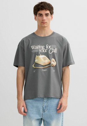 Joven con camiseta gris con gráfico de teléfono rotary vintage y texto "Esperando tu llamada", combinado con vaqueros azul claro.