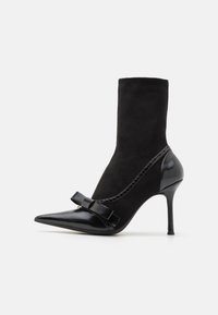 Botine negre cu vârf ascuțit, fabricate din suede și piele. Prezintă un accent sub formă de fundiță și un toc stiletto. Suprafațe netede și texturate.