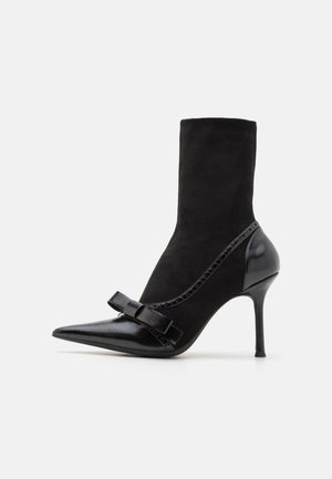 Black ankle boot with a pointed toe, made of suede and leather. Features a bow accent and a stiletto heel. Smooth and textured surfaces.