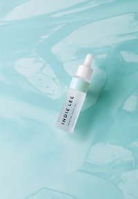 Olej facialny squalan w przezroczystej butelce z kroplomierzem, z białą nakrętką i jasnoniebieską etykietą, umieszczony na miękkozielonym tle.