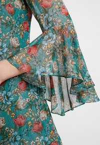 Tissu floral turquoise avec manches cloche orné de fleurs rouges, bleues et beiges ainsi que de fines rayures verticales dorées.