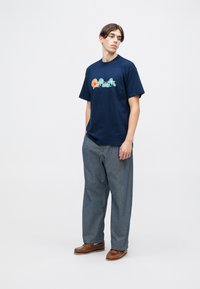 T-shirt in cotone blu navy con logo "Carhartt" multicolore, abbinato a pantaloni di denim grigi larghi e mocassini in pelle marrone.