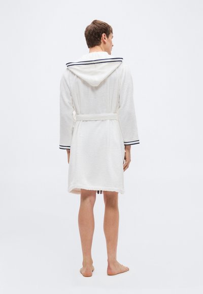 Άνδρας που στέκεται ξυπόλυτος φορώντας λευκή ρόμπα με κουκούλα και μπλε ναυτική διακόσμηση, απεικονίζεται από πίσω σε απλό λευκό φόντο.