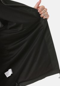 Interno di giacca nera con fodera testurizzata, tessuto esterno liscio e cerniera con ganci laterali. Etichetta di cura visibile in basso.