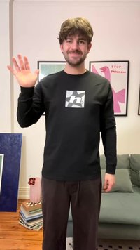 Νεαρός άνδρας με καστανά μαλλιά και γενειάδα κουνάει το χέρι, φοράει μαύρο πουλόβερ με μακριά μανίκια, στέκεται σε σαλόνι με κορνιζαρισμένη τοιχογραφία και στοίβα βιβλίων κοντά.