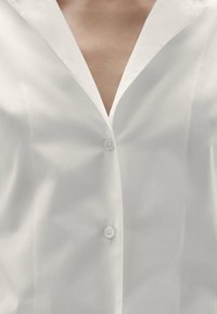 Camicia bianca con bottoni realizzata in un tessuto liscio, caratterizzata da un colletto ampio e tre bottoni visibili sul davanti. Design classico e minimalista.