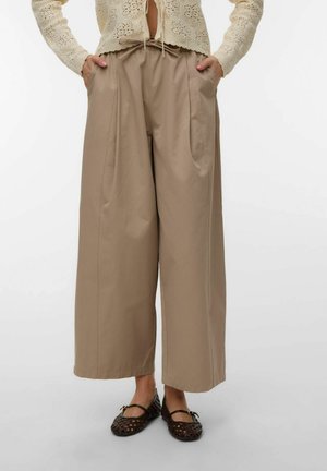 Beżowe spodnie z szerokimi nogawkami, plisami i kieszeniami, noszone z czarnymi plecionymi balerinami Mary Jane oraz kremowym krótkim swetrem z szydełkowym wzorem.