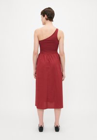 Robe asymétrique à une épaule en rouge foncé, en mélange de coton. Mi-longue avec un corsage ajusté et une jupe fluide. Texture lisse.