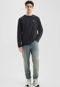Langärmiges, dunkelgraues Hemd mit Rundhalsausschnitt und kleinem Logo auf der Brust, kombiniert mit hellblauen Jeans und Sneakers.