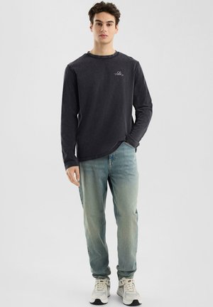 Langsleeve, donkergrijze shirt met een ronde hals en een klein logo op de borst, gecombineerd met lichtblauwe denim jeans en sneakers.