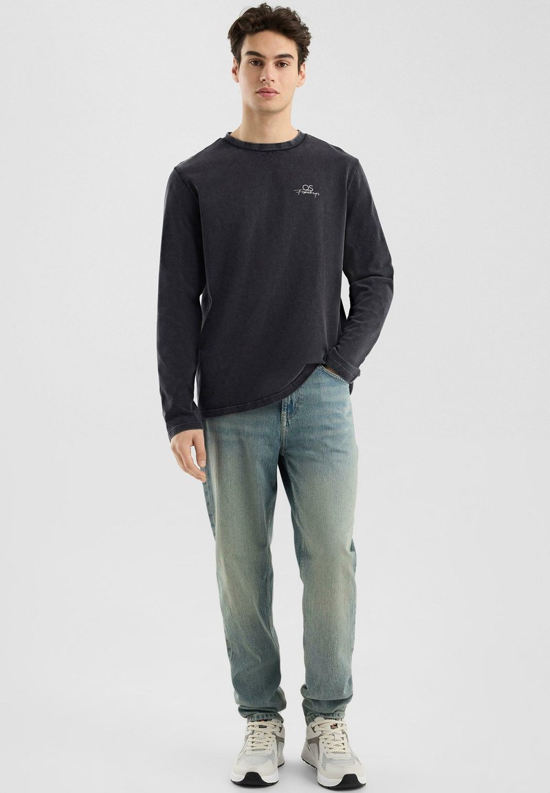 Langärmiges, dunkelgraues Hemd mit Rundhalsausschnitt und kleinem Logo auf der Brust, kombiniert mit hellblauen Jeans und Sneakers.