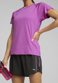 Lekka fioletowa sportowa koszulka z krótkimi rękawami, zestawiona z czarnymi szortami z różowym akcentem oraz różową butelką na wodę.