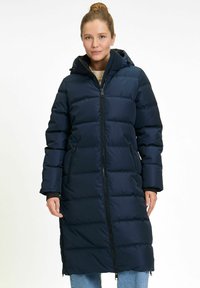 Vrouw in een lange, marineblauwe, gewatteerde winterjas met capuchon, voorrits en ritszakken, staand tegen een effen achtergrond.