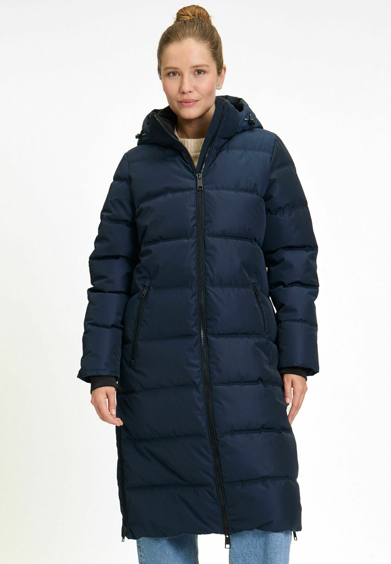 Vrouw in een lange, marineblauwe, gewatteerde winterjas met capuchon, voorrits en ritszakken, staand tegen een effen achtergrond.
