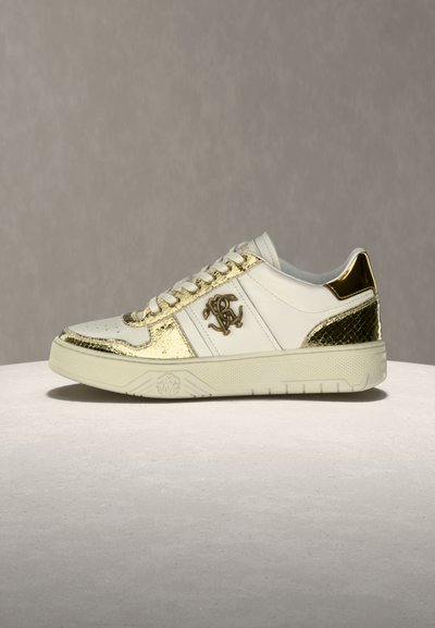 Baskets basses blanches avec accents métalliques dorés, lacets et logo décoratif sur le côté, présentées sur une surface claire.