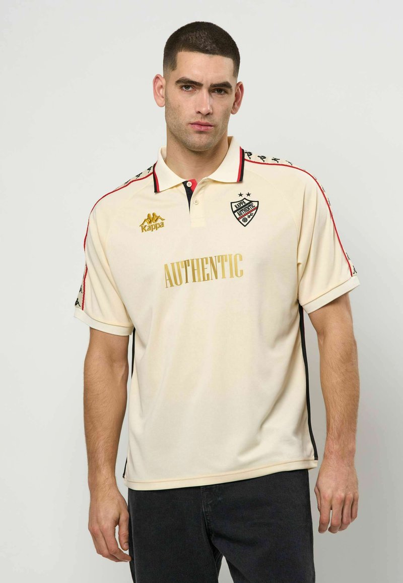 Μπεζ πόλο μπλουζάκι με μαύρες και κόκκινες λεπτομέρειες. Φέρει το λογότυπο "Kappa" και τη λέξη "AUTHENTIC" σε χρυσό χρώμα. Κοντά μανίκια και κλασικός γιακάς.