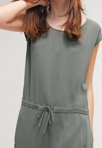 Mujer vestida con un mono sin mangas gris y cintura ajustable, posando con una mano detrás de la cabeza sobre un fondo liso.