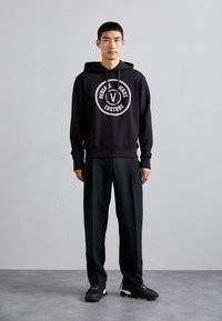 Felpa nera con cappuccio caratterizzata da un grande logo bianco con design a catena, abbinata a pantaloni neri a gamba larga e scarpe sportive nere.