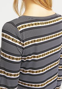 Kvinna som bär en svart långärmad topp med horisontella vita prickränder och ett mönster av guldkedjor över ryggen och ärmarna.