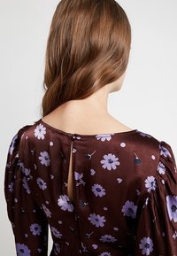 Brun blommig klänning med lila blommor, puffärmar och en nyckelhålsdetalj på ryggen. Slät satinstruktur med en lätt lyster.