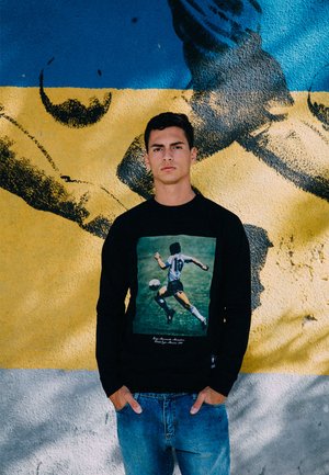 Zwarte sweatshirt met een grafisch ontwerp van een voetballer in actie. De speler draagt een wit shirt met zwarte shorts. Daaronder zijn blauwe spijkerbroeken.