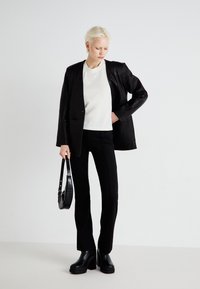 Crna kožna blazer jakna preko bijele majice, u kombinaciji s crnim uskim hlačama i debelim crnim platformskim čizmama. Model drži malu crnu torbicu.