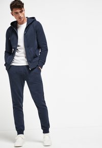 Marineblaue Kapuzenjacke und Jogginghose aus weichem Stoff. Mit vorderen Taschen, Reißverschluss und ribbeligem Bündchen. Kombiniert mit weißen Sneakers.