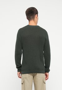 Mann mit einem dunkelgrünen Rippstrickpullover und beigen Cargo-Shorts, der mit dem Rücken zu einer schlichten weißen Wand steht.