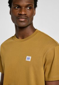 Braunes Baumwoll-T-Shirt mit kurzen Ärmeln und Rundhalsausschnitt. Auf der Vorderseite befindet sich ein kleines blau-weißes Markenpatch. Glatte Textur.