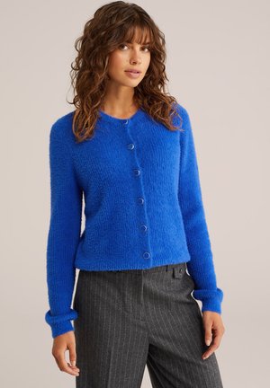 Helderblauwe fuzzy cardigan met ronde zilverkleurige knopen, lange mouwen en geribde manchetten. Gekozen boven een grijze gestreepte pantalon.