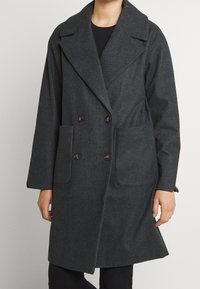 Mörkgrå dubbelknäppt kappa i herringbone-tyg. Har stora framfickor och breda slag, med en avslappnad passform.