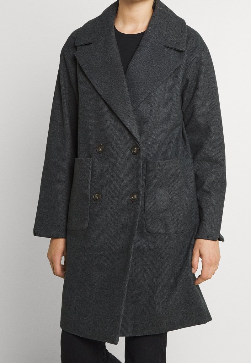 Mörkgrå dubbelknäppt kappa i herringbone-tyg. Har stora framfickor och breda slag, med en avslappnad passform.