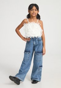 Top corto bianco con volant e spalline sottili abbinato a jeans cargo blu a gamba larga con molte tasche. Calzature nere completano il look.