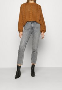 Mujer vestida con un suéter de punto trenzado color óxido, jeans de tiro alto en gris deslavado y botas negras hasta el tobillo, contra un fondo claro.