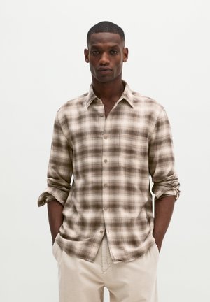 Homme portant une chemise à carreaux marron et beige boutonnée avec les manches retroussées et un pantalon en velours côtelé clair, mains dans les poches.