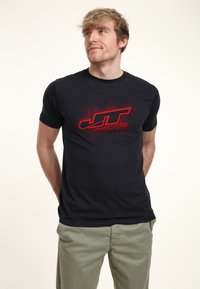 Ung mand med blond hår iført en sort JT Racing t-shirt og lysegrønne bukser, stående med hænderne bag ryggen.