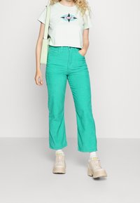 Camiseta cortada verde menta con un logo gráfico, combinada con pantalones de pana teal de tiro alto y con estrías, y zapatillas beige gruesas.