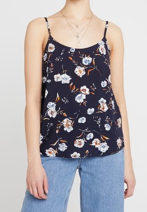 Kobieta w granatowym kwiecistym topie na cienkich ramiączkach, w jasnoniebieskich dżinsach i warstwowych złotych naszyjnikach.