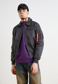 Black bomber jacket with a zip front, ribbed cuffs, and a patch on the chest. Features a red strap on the sleeve and is paired with a purple sweatshirt.