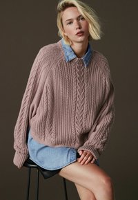 Üks suur roosa kaelusnööbiga kampsun, mille all on kerge denimpusa, ribidega mansettide ja tekstuuriga mustriga.