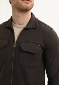 Giacca zip nera in tonalità marrone scuro con colletto a camicia, due tasche sul petto e una texture morbida. Indossata sopra una camicia beige.